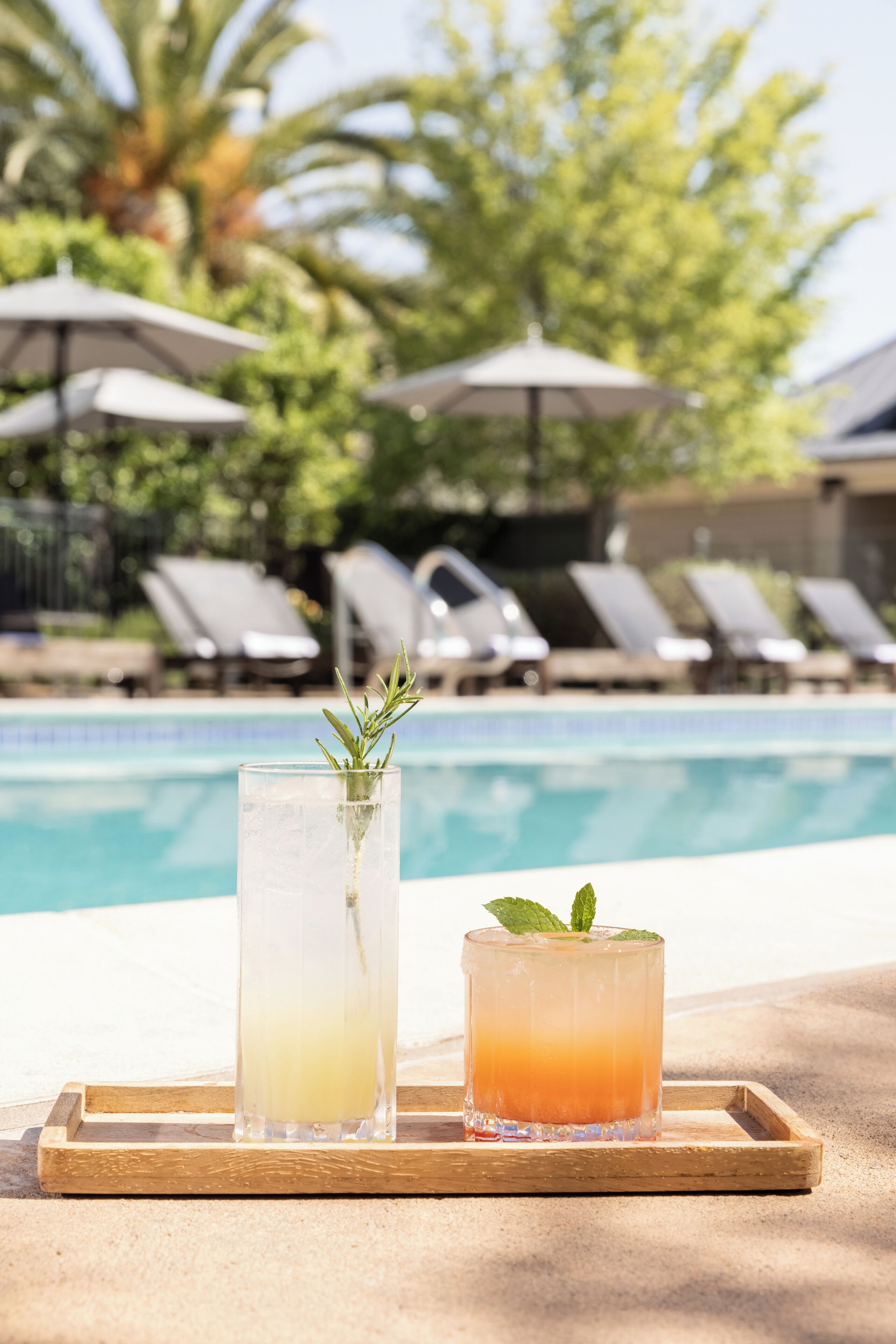 A poolside setting with two drinks on a tray, a pool, and lounge chairs in the background.