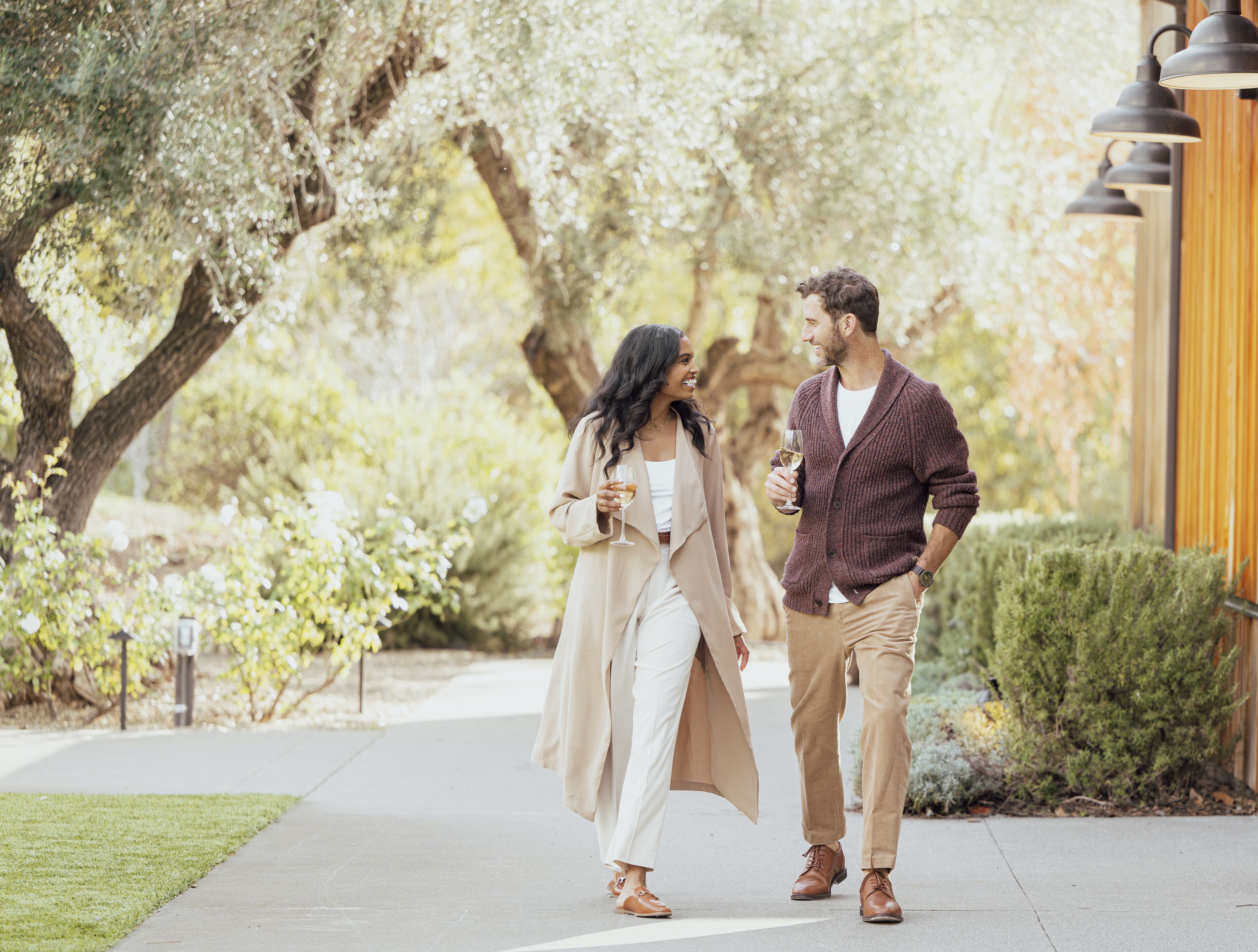 A couple walking on a sidewalk with trees and a building in the background.