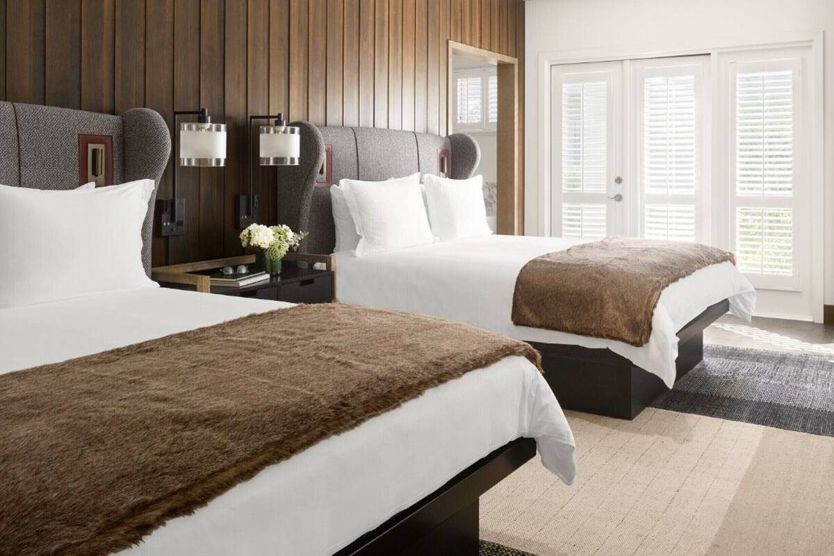 A modern hotel room with two beds, wooden paneling on the walls, a large mirror, and a view of the outdoors through the door.