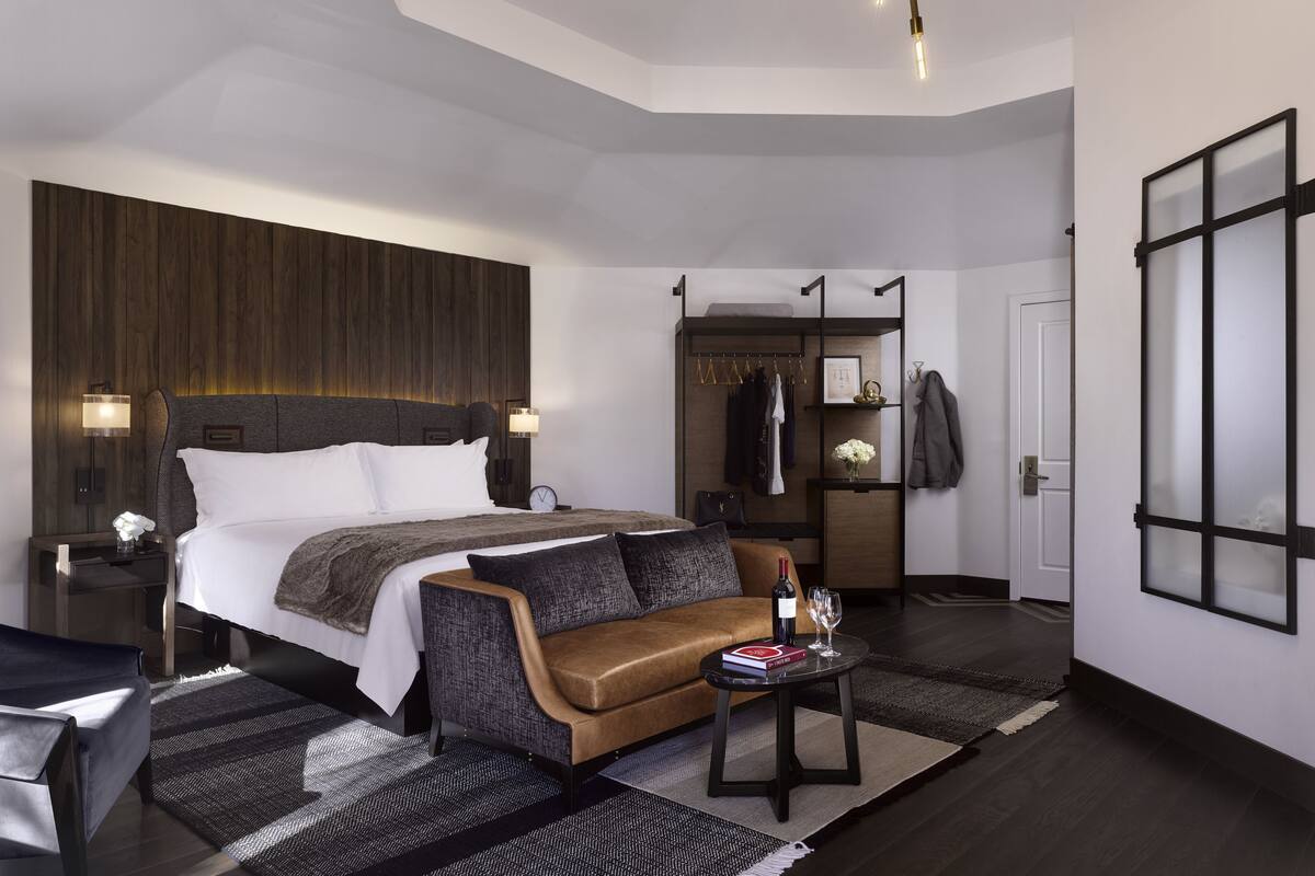 A modern hotel room with a large bed, a sofa, a small table with a book and wine glass, and a wardrobe with hanging clothes.