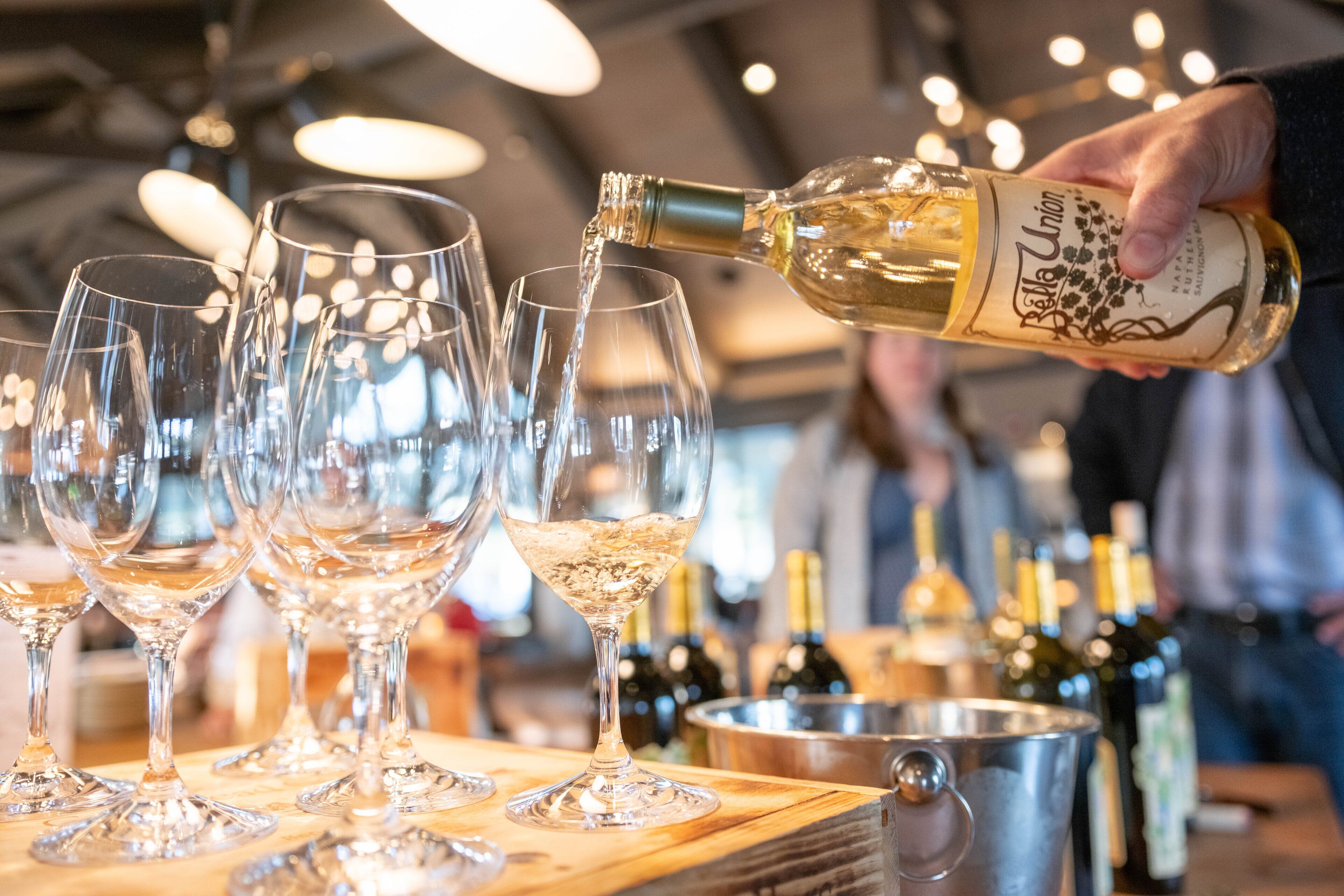 A person pouring wine into glasses on a bar counter with bottles and a bucket in the background.