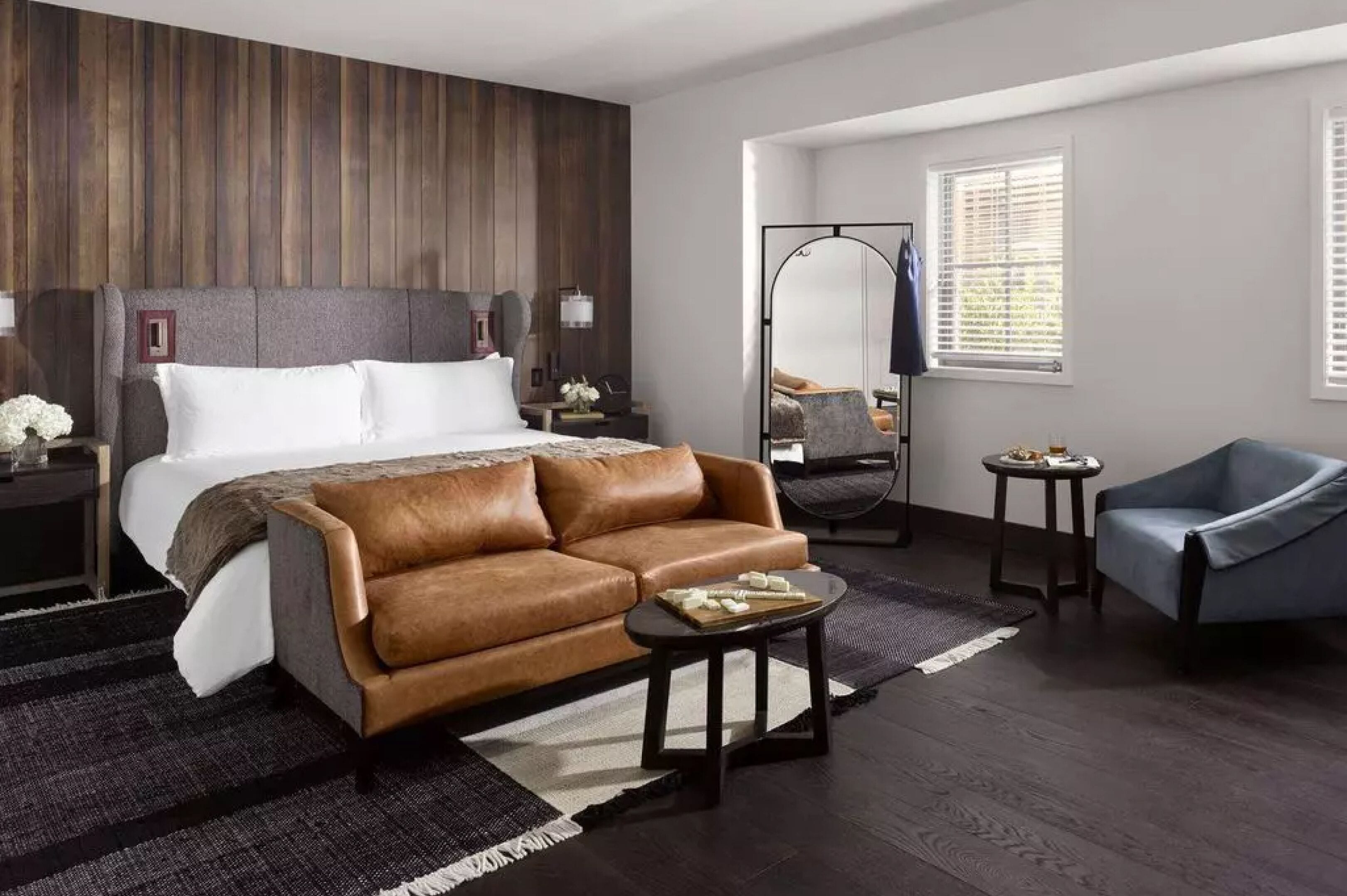 Una habitación de hotel moderna con una cama grande, un sofá de cuero marrón, una mesita redonda pequeña y un sillón azul.