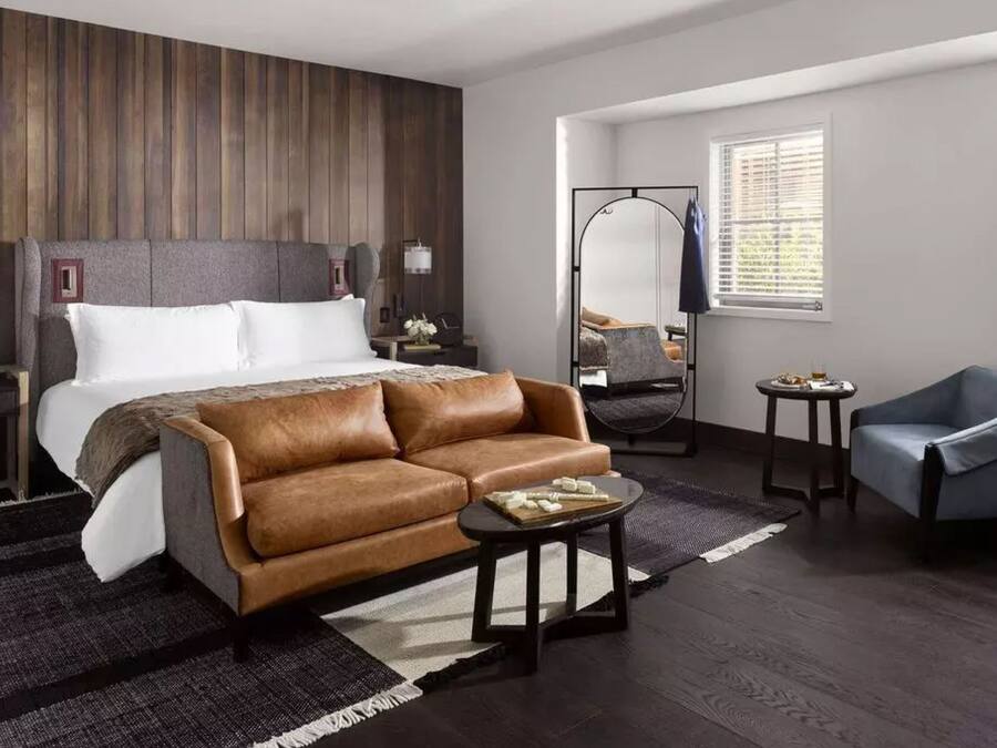 Una habitación de hotel moderna con una cama grande, un sofá de cuero marrón, una mesita redonda pequeña y un sillón azul.
