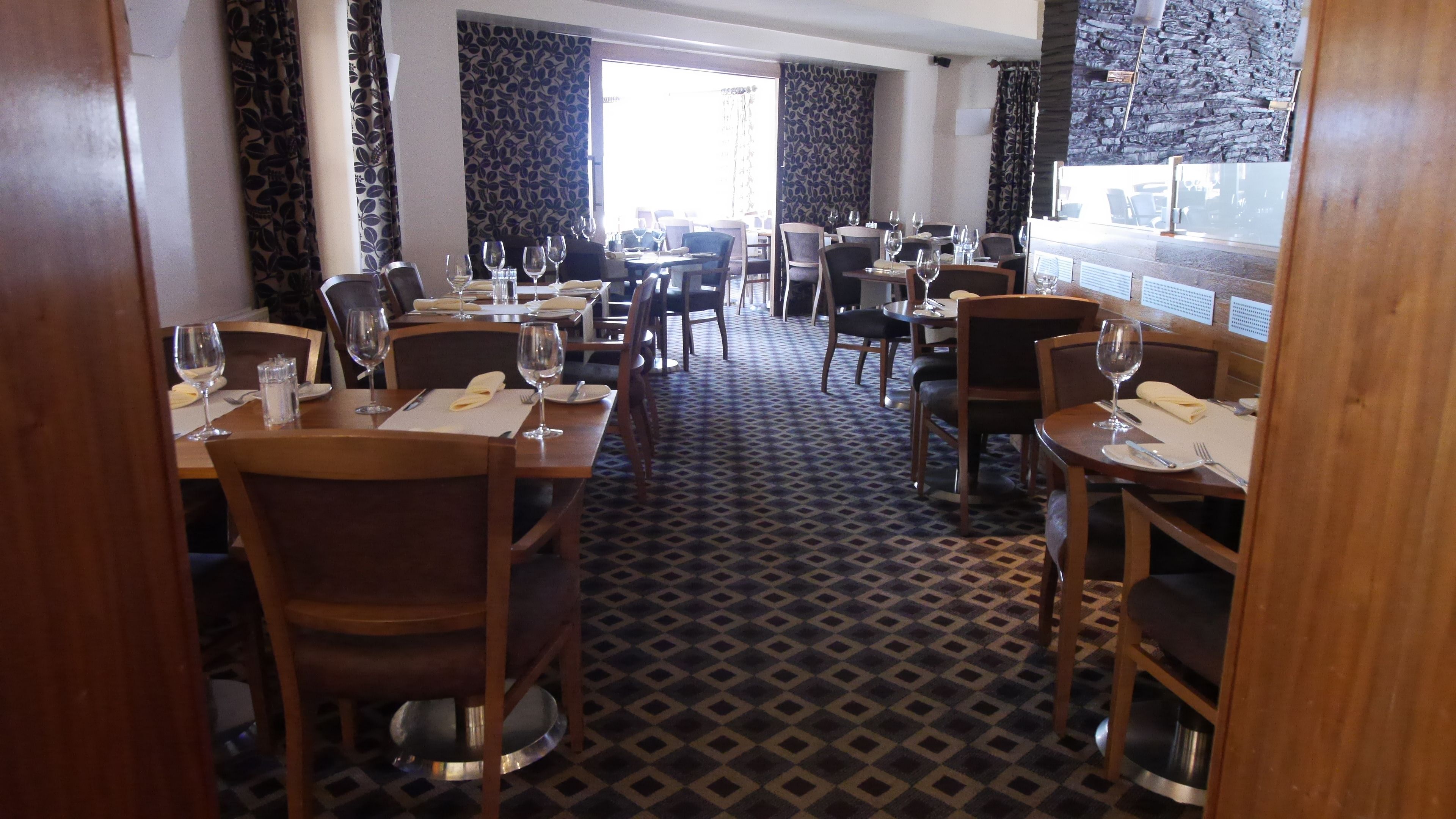 A restaurant with wooden tables and chairs, a patterned carpet, and a view through large windows.