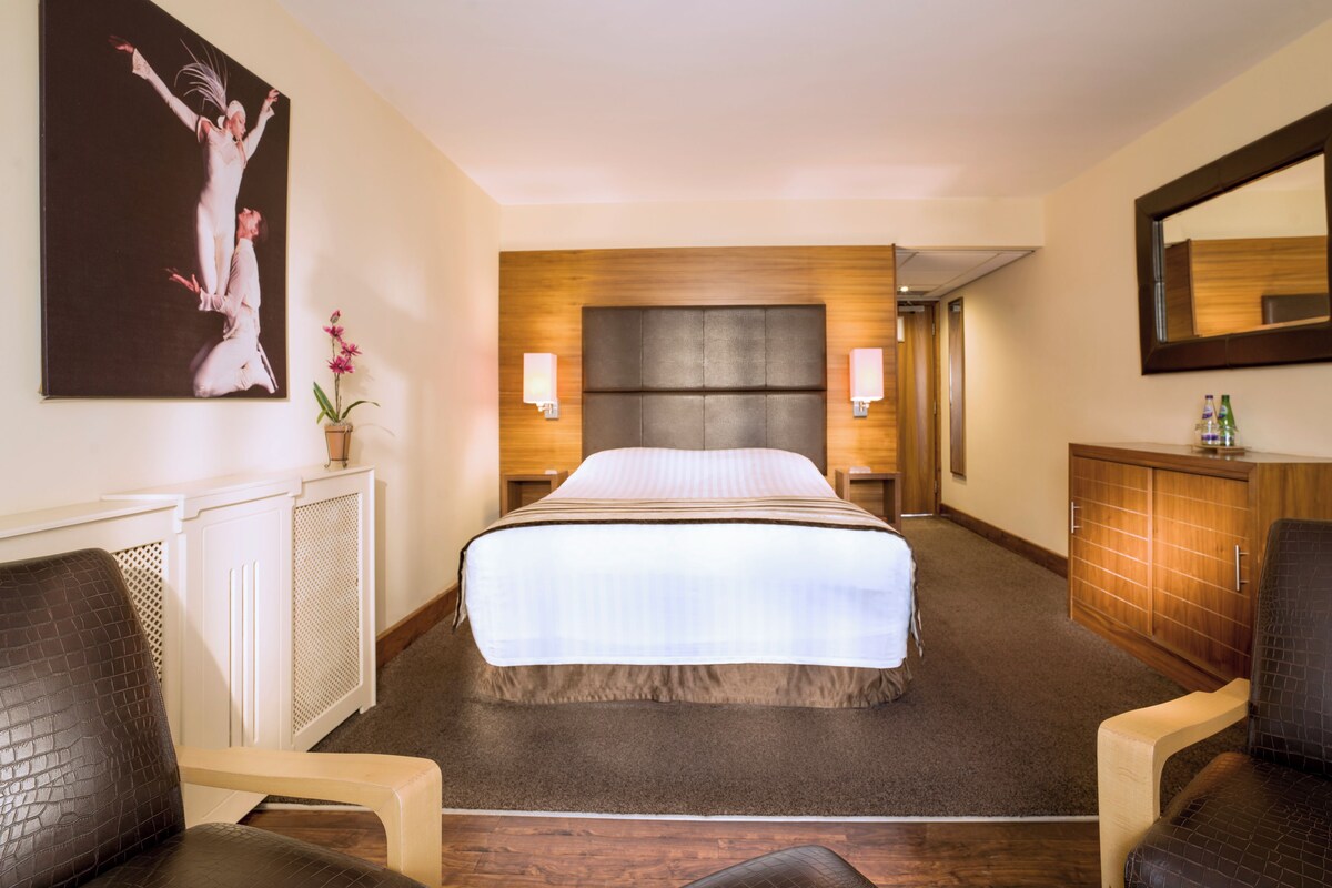 A hotel room with a large bed, two bedside lamps, a wooden headboard, a mirror, a wooden cabinet, and a painting on the wall.