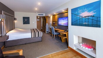 Een hotelkamer met een bed, bureau, stoel, televisie en een schilderij van een brug.