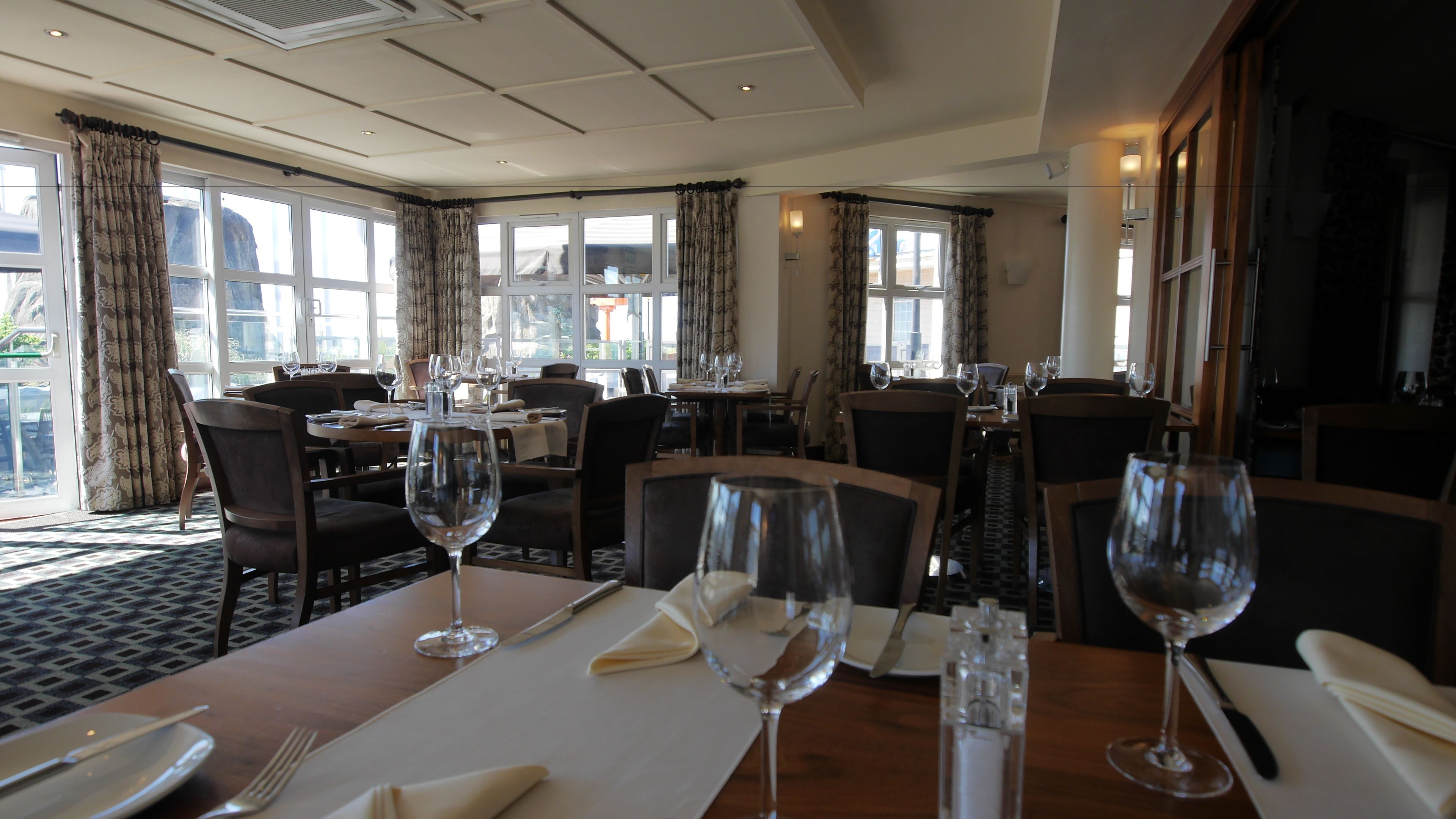 A restaurant with multiple tables, each set with wine glasses, cutlery, and napkins. Large windows with curtains allow natural light to enter.