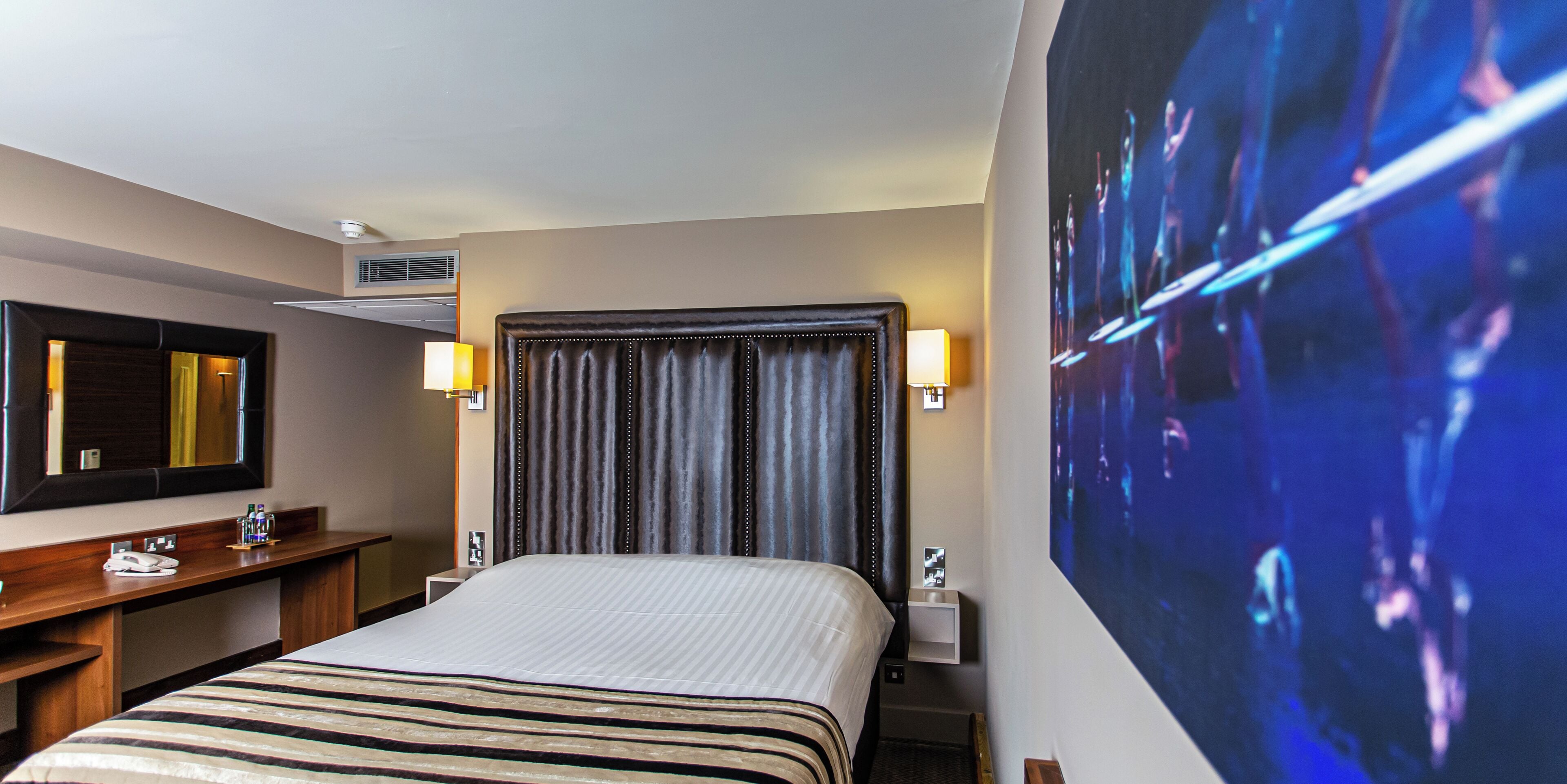 A hotel room with a large bed, a desk with a telephone, a mirror, and a wall-mounted TV.