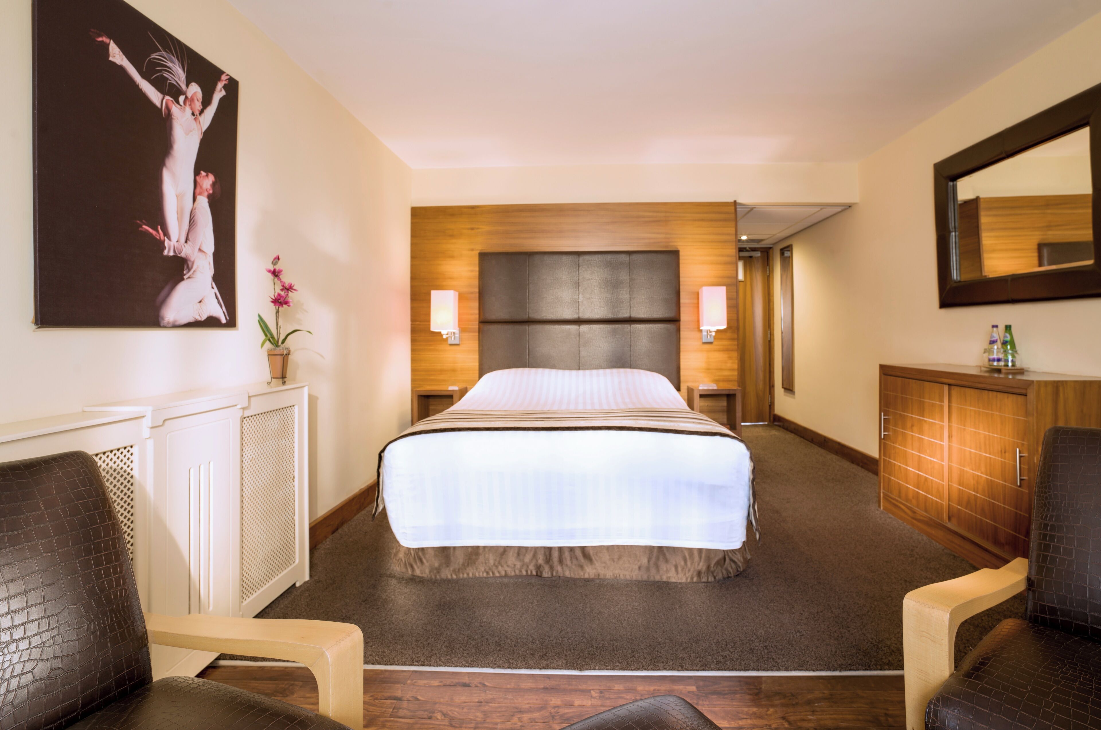 A hotel room with a large bed, two bedside lamps, a wooden headboard, a mirror, a wooden cabinet, and a painting on the wall.
