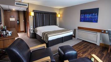 A hotel room with a large bed, a desk with a chair, a small table, a radiator, and a wall-mounted TV.