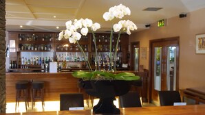 Una zona de bar con una jarra negra de orquídeas blancas sobre la barra, sillas de madera y estanterías llenas de diferentes botellas.