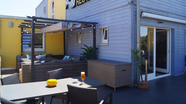 An outdoor seating area with a pergola, wicker furniture, and a glass table with a yellow bowl and a glass of beer.