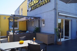 Un espacio de estar al aire libre con una pérgola, mobiliario de mimbre y una mesa de vidrio con un tazón amarillo y una cerveza.
