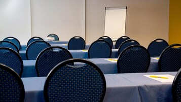 Un salón de conferencias con filas de mesas y sillas, una pantalla de proyector y una pizarra.