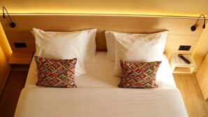 A neatly made bed with two white pillows and two patterned pillows, a bedside table with a remote control, and a wall-mounted lamp.