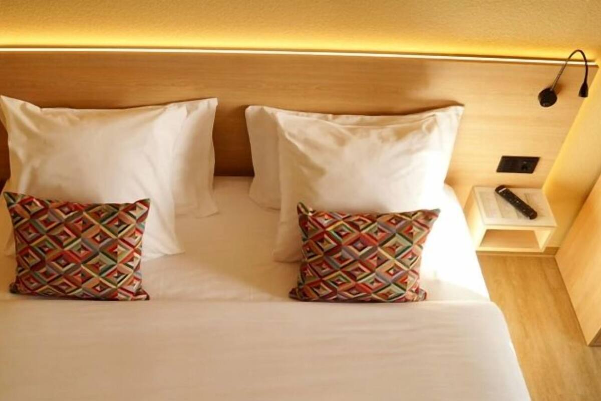 A neatly made bed with two white pillows and two patterned pillows, a bedside table with a remote control, and a wall-mounted lamp.