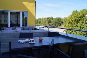 A balcony with a sofa, table, and chairs, overlooking a parking area and trees.