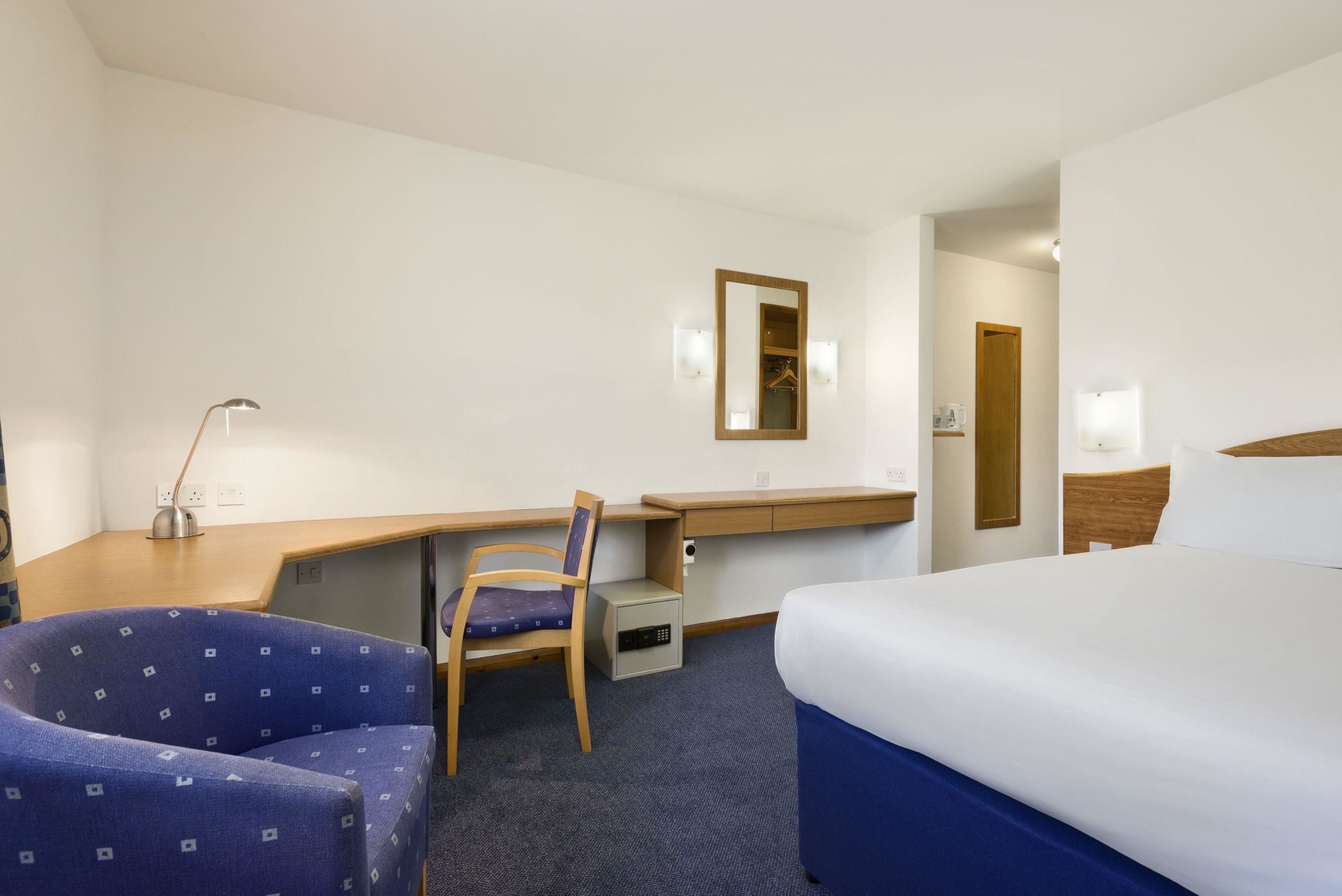 A hotel room with a bed, a desk, a chair, a small cabinet, and a mirror.