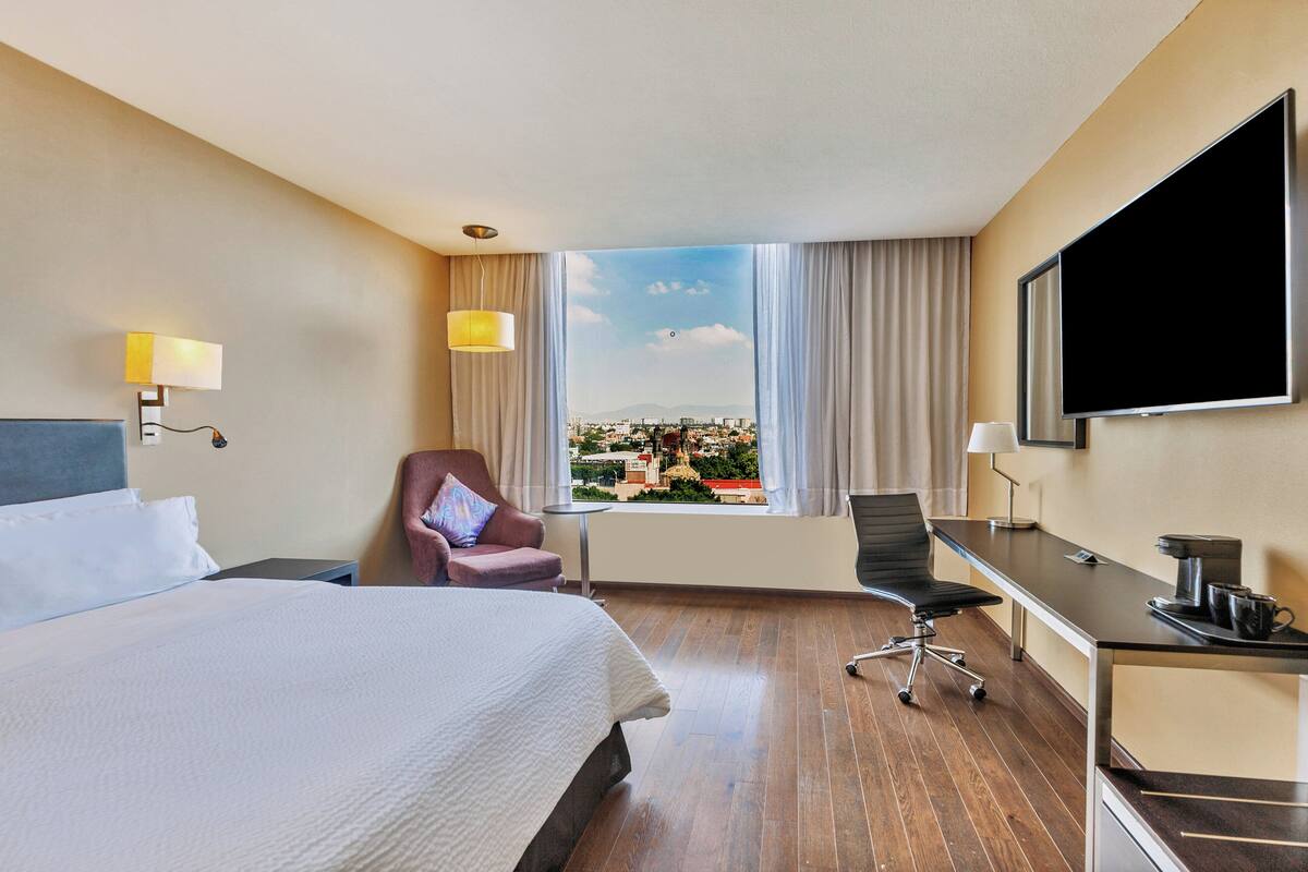 A hotel room with a large bed, a desk, a chair, a TV, and a window with a city view.