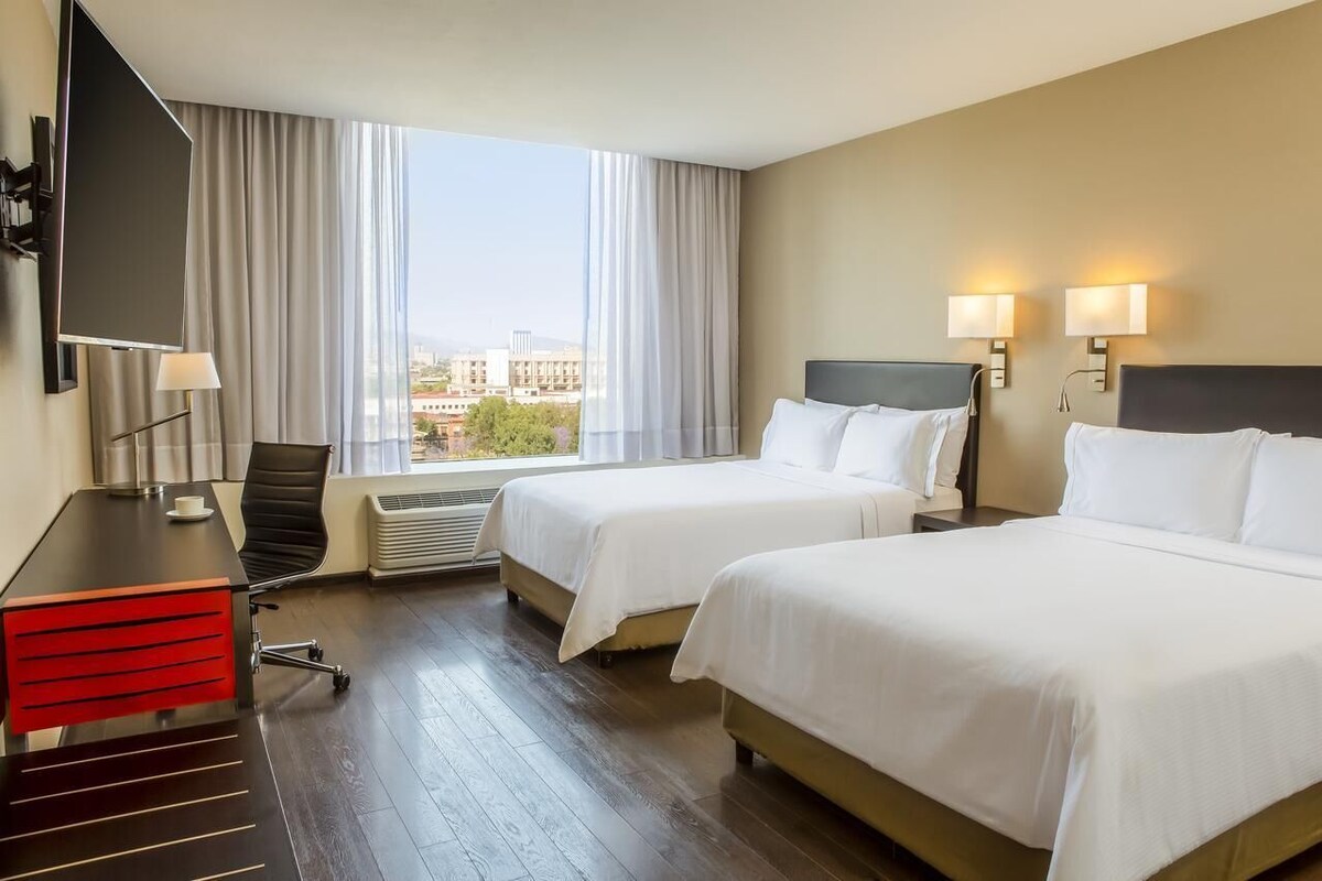 Habitación de hotel con dos camas, un escritorio con silla, televisión y vistas de la ciudad.