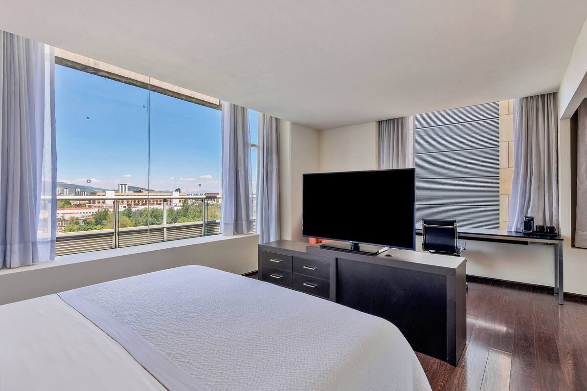A hotel room with a large bed, a TV, a desk, and a view of the city.
