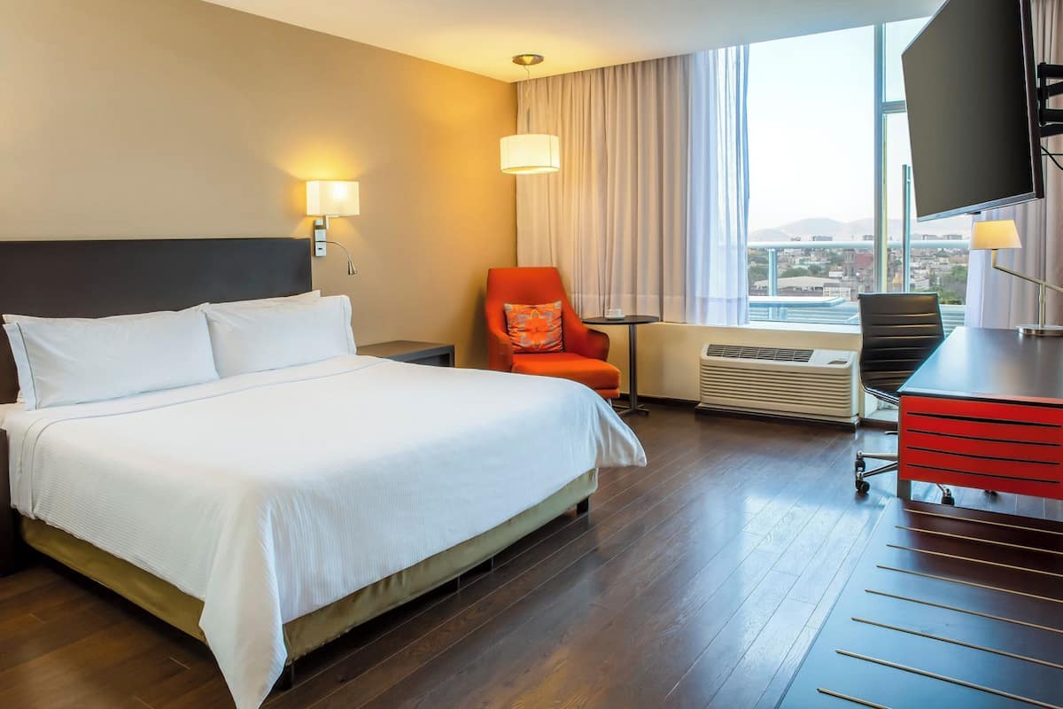 A hotel room with a bed, a desk, an orange chair, a TV, and a large window with a city view.