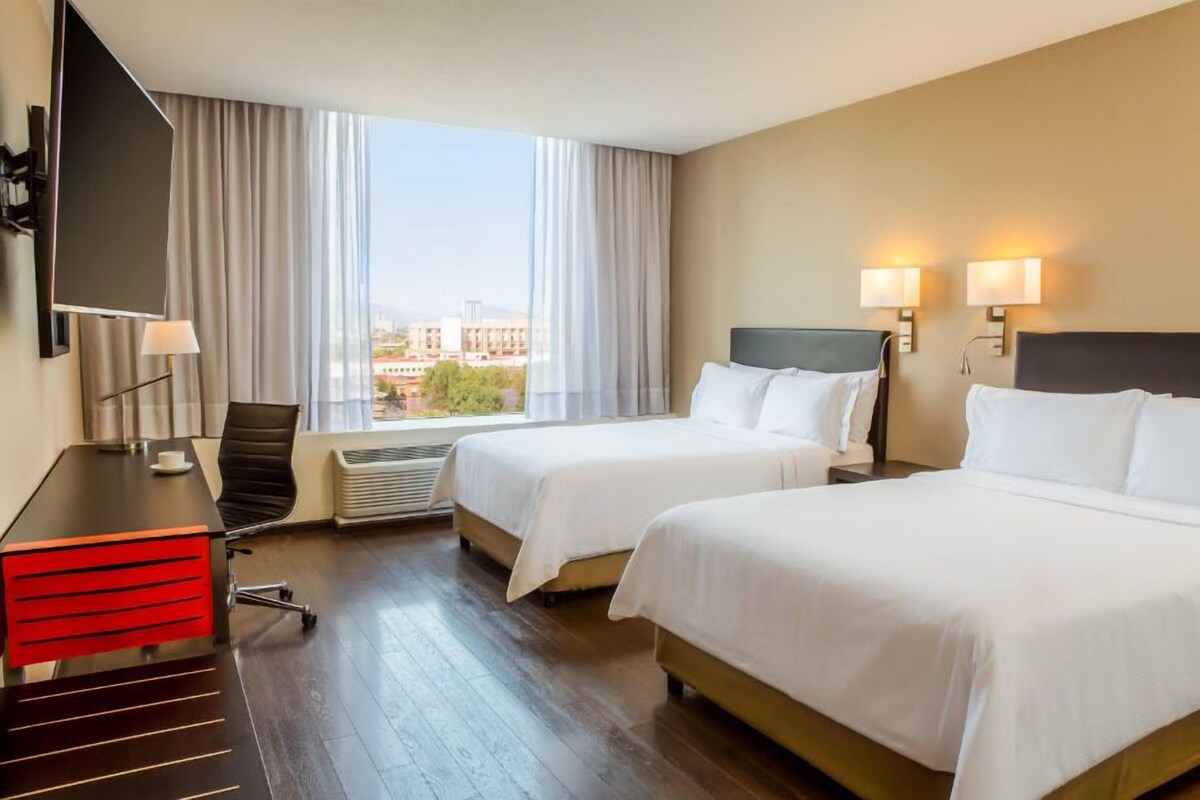 Habitación de hotel con dos camas, un escritorio con silla, televisor y una ventana grande con cortinas.