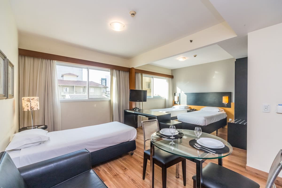 Quarto de hotel com duas camas, área de refeições com mesa de vidro e vista para edifícios.
