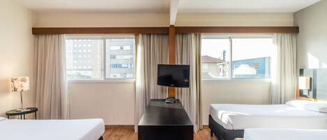 A hotel room with two beds, a TV on a stand, a lamp, and large windows with curtains.
