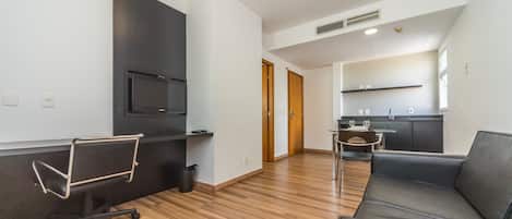 Una sala de estar moderna con un sofá de cuero, una silla de oficina negra, un televisor montado en la pared y una cocina al fondo.