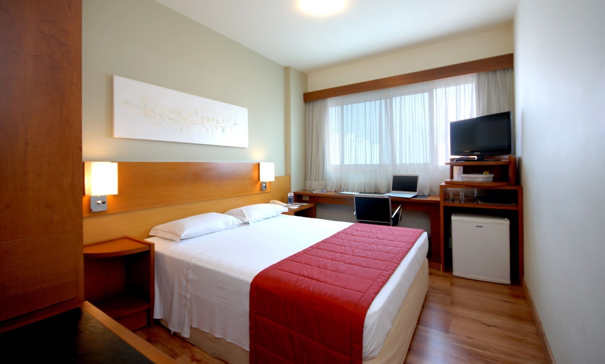 A hotel room with a bed, a desk with a computer, a television, and a small refrigerator.