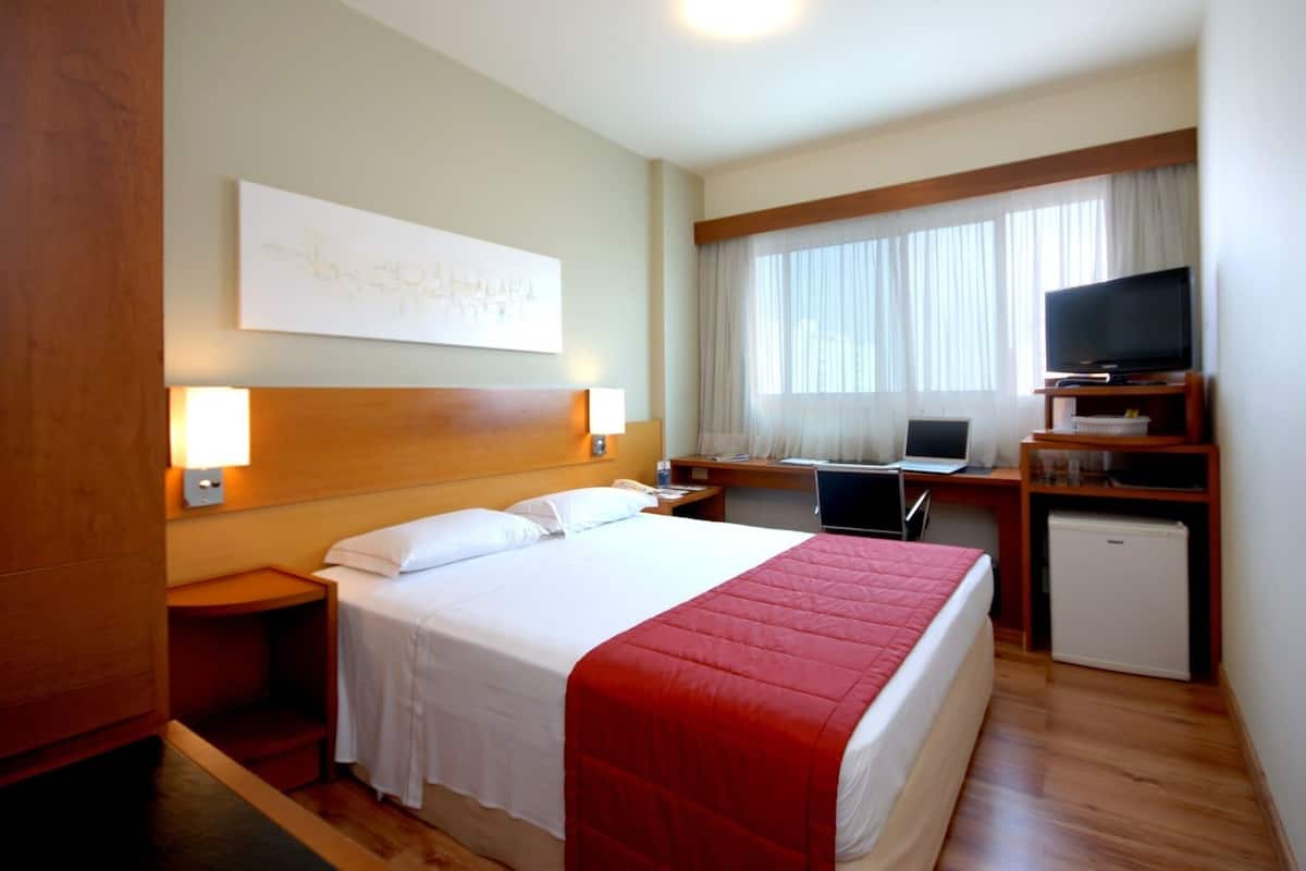 A hotel room with a bed, a desk with a computer, a television, and a small refrigerator.