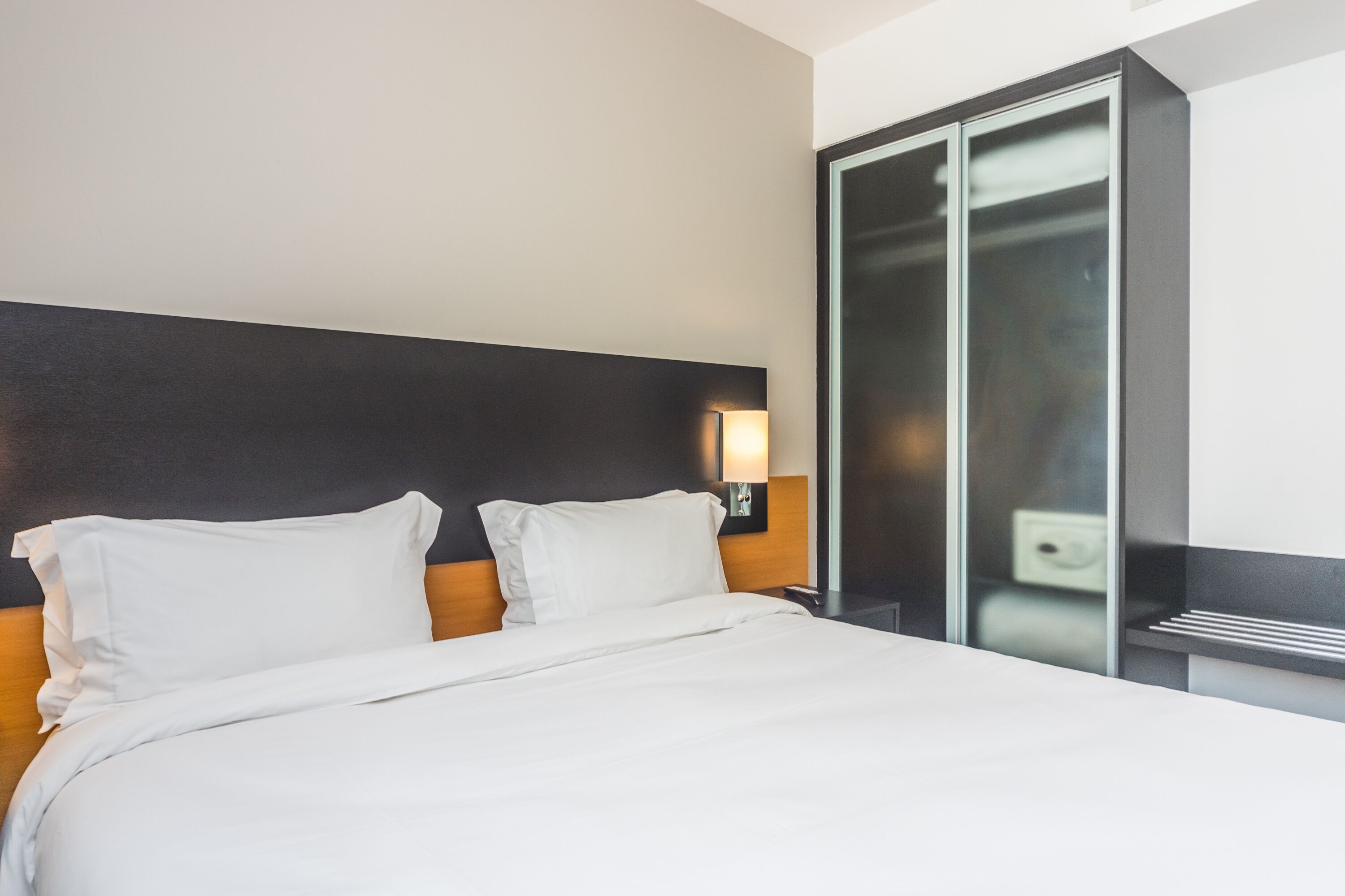 A modern hotel room with a large bed, a wooden headboard, and a glass partition wall.
