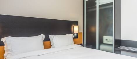 A modern hotel room with a large bed, a wooden headboard, and a glass partition wall.