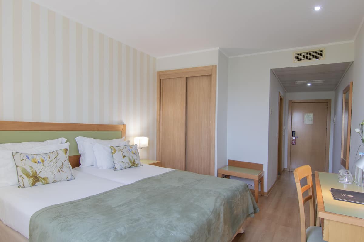 A hotel room with a large bed, a wooden wardrobe, a desk with a chair, and a small table with a lamp.