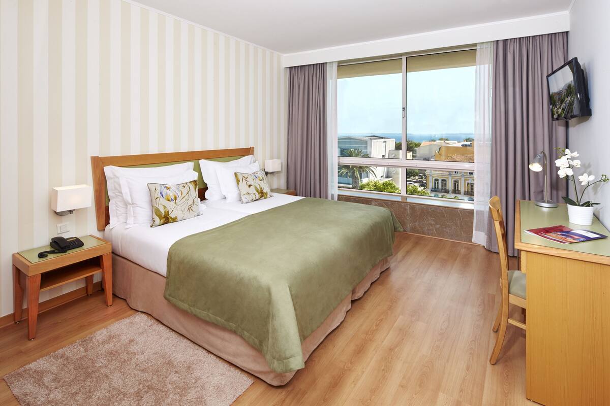A hotel room with a large bed, a desk, a chair, a TV, and a balcony with a view.