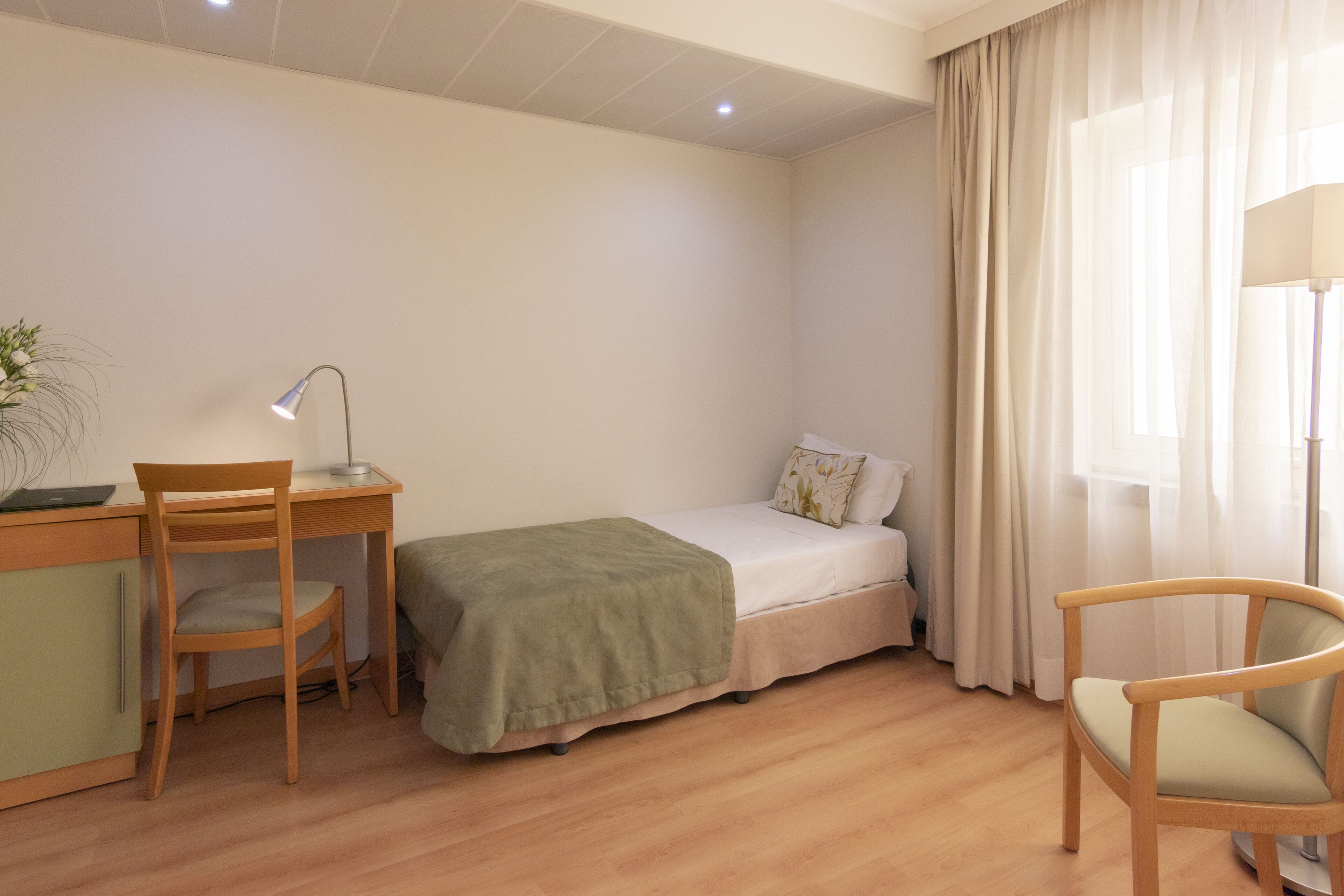 A small, neatly arranged room with a bed, a desk with a chair, a lamp, and a window with curtains.