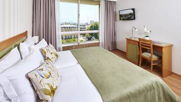 A hotel room with a bed, a desk, a chair, a lamp, and a view of buildings through the window.