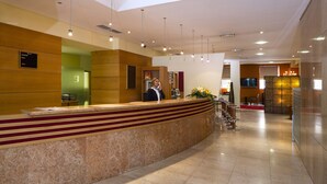 Zona de recepción de un hotel con mostrador de mármol, una recepcionista y una pantalla de televisión.