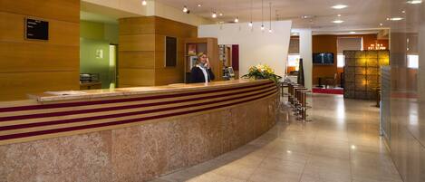Een hotel receptie met een marmeren balie, een receptionist en een televisiescherm.
