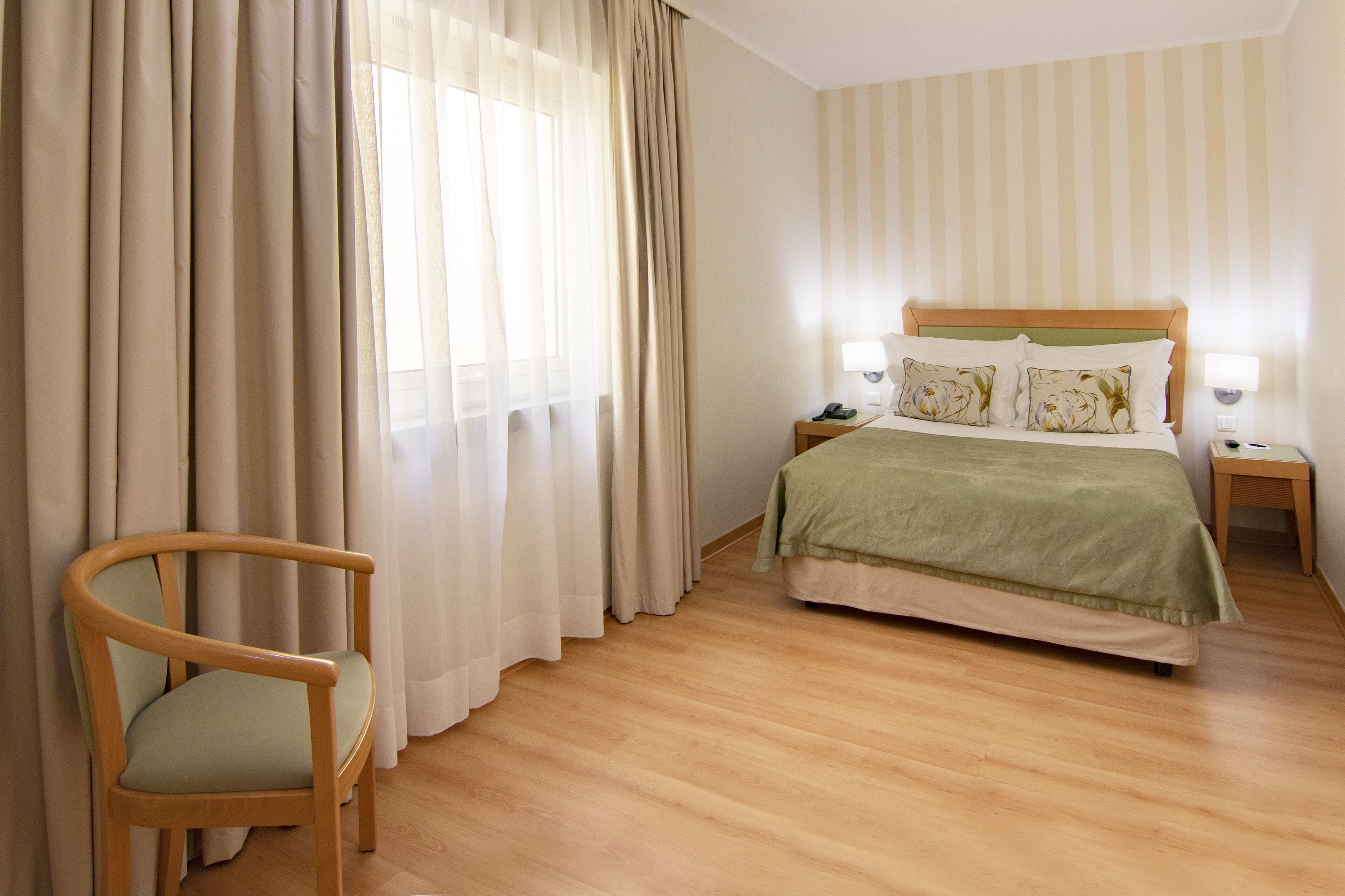 A hotel room with a wooden floor, a bed with a green bedspread, a wooden chair, and a nightstand with a lamp.