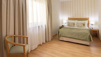 A hotel room with a wooden floor, a bed with a green bedspread, a wooden chair, and a nightstand with a lamp.