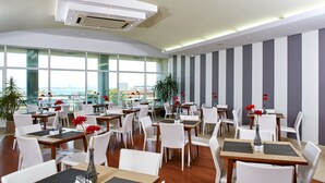 Un comedor de hotel moderno con varias mesas y sillas, amplias ventanas que ofrecen vistas de la ciudad y un diseño de pared a rayas.