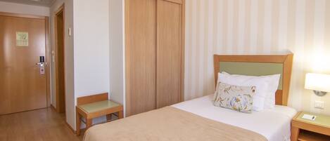 Habitación de hotel con una cama, un armario de madera, una mesita pequeña y una mesita de noche con lámpara.