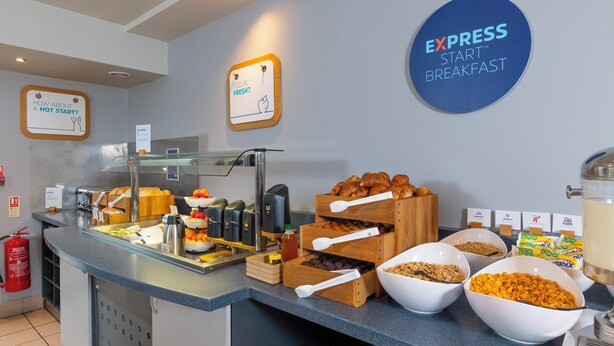 A breakfast buffet area with various food items, a coffee machine, and a sign that reads "EXPRESS START BREAKFAST".