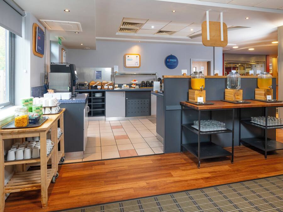 A modern breakfast area with a coffee machine, a counter with a sign reading "Express Breakfast," and a seating area with a wooden table and chairs.