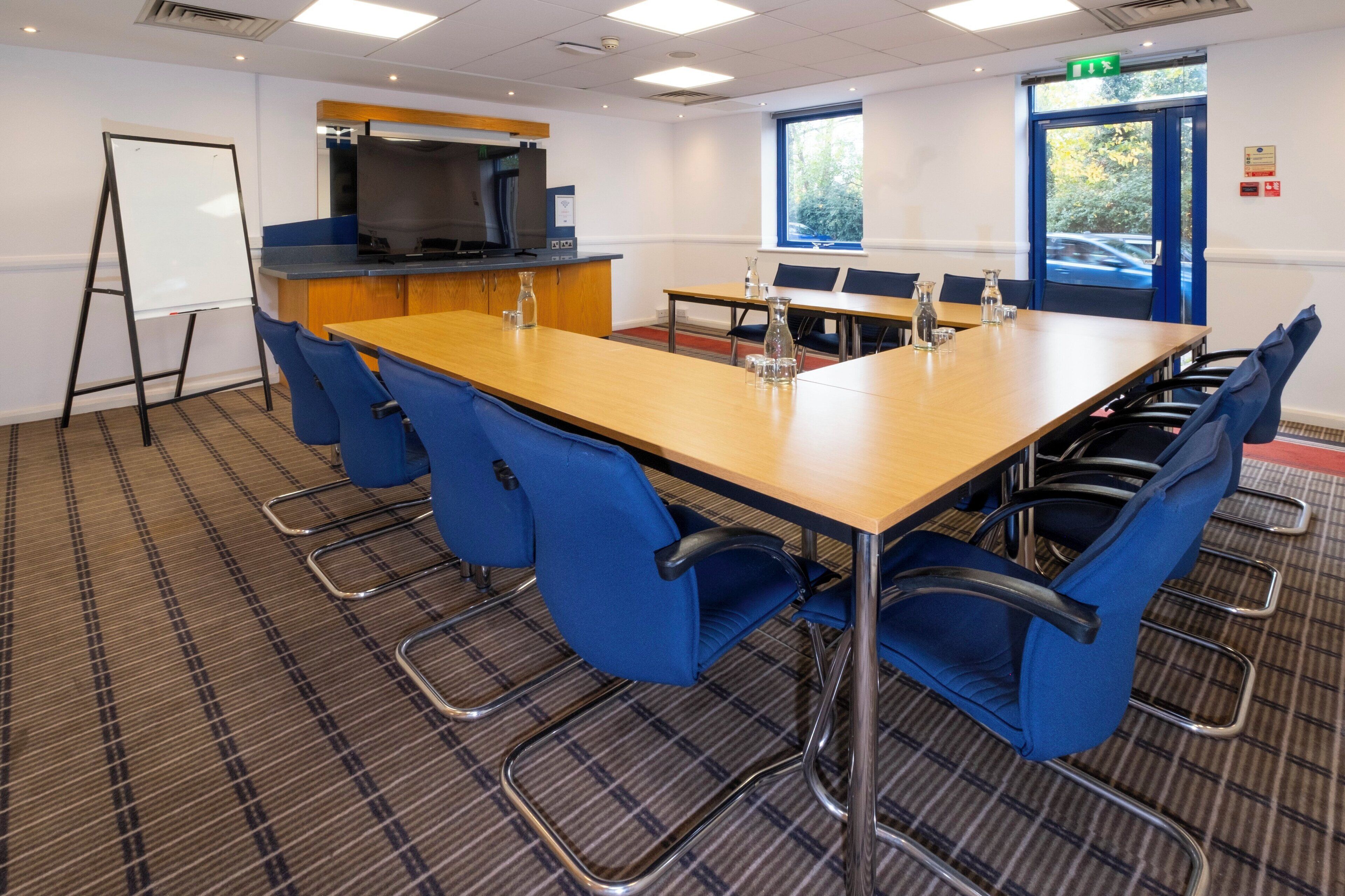 A conference room with a long table, blue chairs, a whiteboard, a television, and a window with a view of trees.