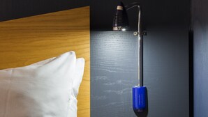 A neatly made bed with white linens, a bedside table with a wine glass and a book, and a wall-mounted lamp with a blue base.