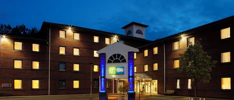 Un hotel de varios pisos con una entrada central que presenta un arco iluminado en azul y una torre del reloj.