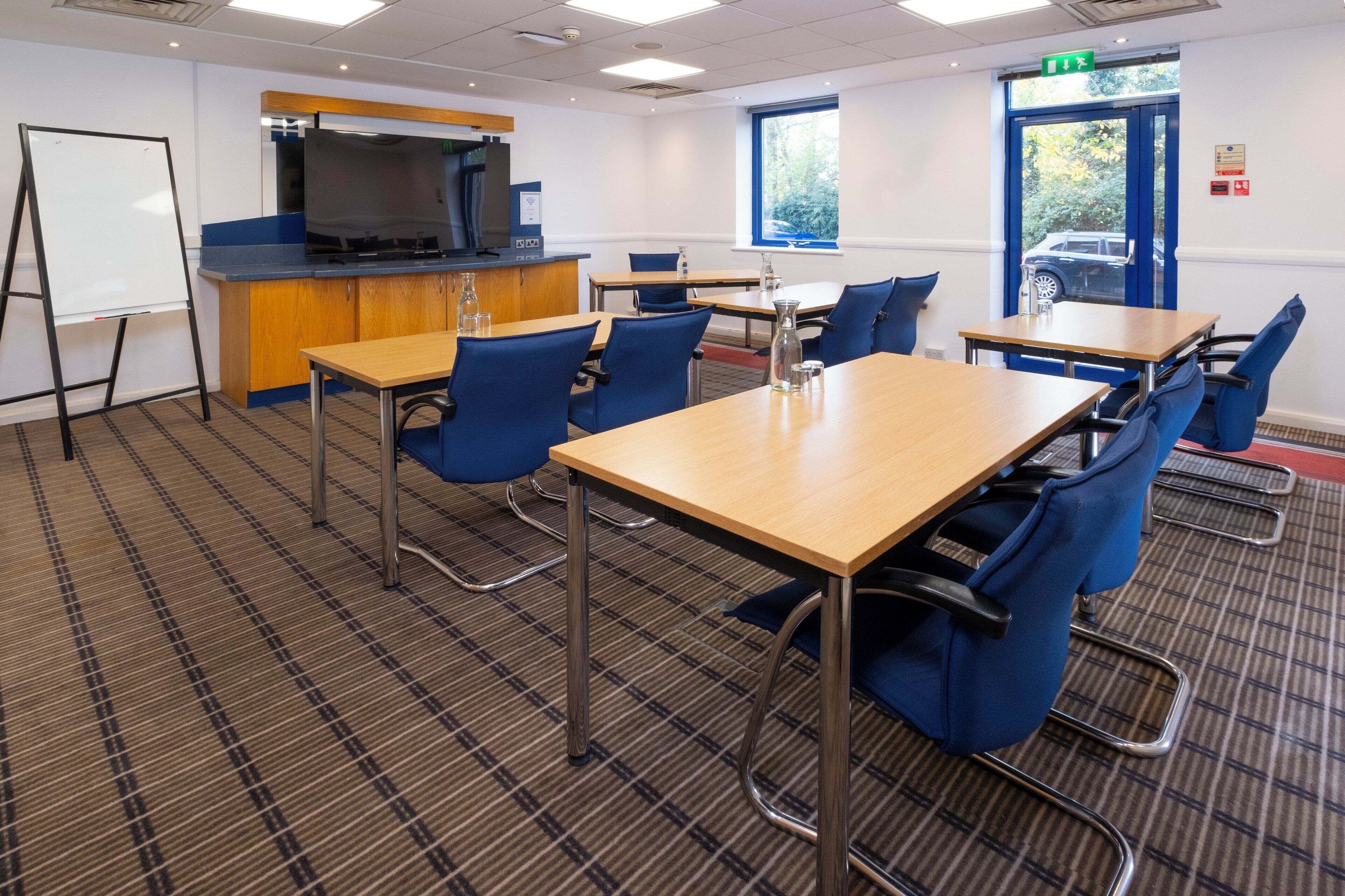 A conference room with a large table, blue chairs, a whiteboard, a kitchenette, and a window with a view of a parked car.